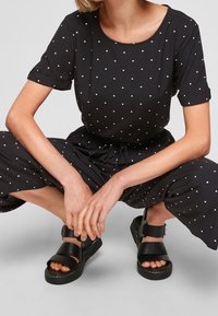 Combinaison noire à pois blancs avec manches courtes, taille élastique et coupe décontractée ; associée à des sandales noires épaisses avec larges brides.