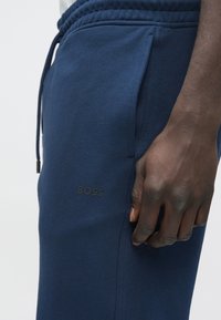Donkere hand rust naast marineblauwe joggingbroek met zwart "BOSS"-logo en zichtbare trekkoordjes tegen een lichte achtergrond.