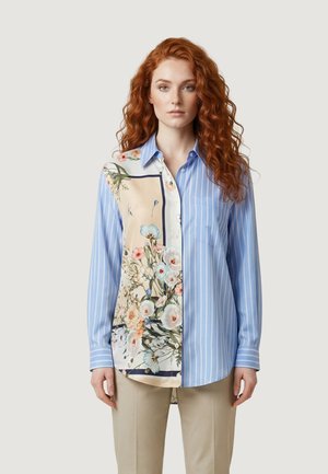 Femme aux cheveux roux bouclés portant une chemise à manches longues avec des rayures verticales bleues et un panneau à motifs floraux, accompagnée d'un pantalon beige.