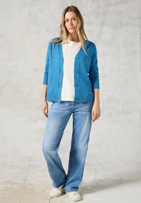 Ljust blå cardigan med knappstängning och ribbade ärmar, bärs över en vit skjorta och ljusblå jeans. Modellen står mot en neutral bakgrund.