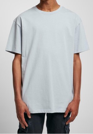 Mann in einem schlichten, hellgrauen, oversized Kurzarm-T-Shirt und schwarzen Cargo-Hosen, der mit entspannt hängenden Händen dasteht.