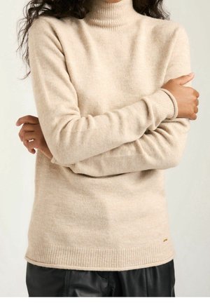Personne portant un pull à col roulé beige à manches longues et un pantalon noir, les bras croisés, sur un fond clair uni.