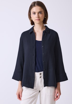 Dark navy button-up shirt made of textured fabric with a collar and wide sleeves, paired with a navy top and white pants.