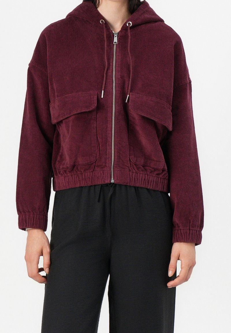 Femme portant une veste courte en velours côtelé bordeaux avec capuche, fermeture éclair frontale et grandes poches à rabat, associée à un pantalon noir ample.