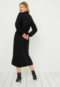 Czarna sukienka z długim rękawem, wysokim kołnierzem, plisami z tyłu i dopasowanym pasem. Nosi się z czarnymi botkami za kostkę na obcasie.