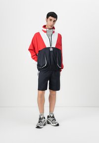 Farveblokeret vindjakke i rød, hvid og marineblå med lynlås og forlommer, parret med marineblå shorts og sorte og sølv sneakers.