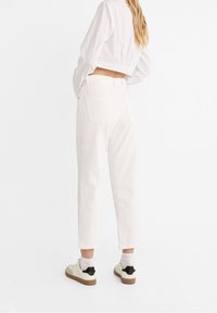 Bílé bavlněné kalhoty s přímým střihem a pěti kapsami, viděné zezadu. Nošeny s krátkým bílým topem a béžovými teniskami.