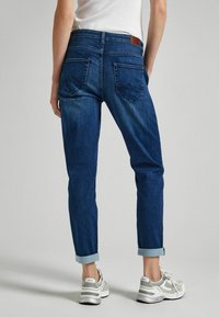 Jeans de mezclilla azul con un corte ajustado, con dobladillos enrollados, dos bolsillos traseros y un parche de cuero marrón en la cintura. Combinados con zapatillas.