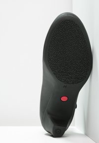 Semelle de chaussure noire présentant un motif floral complexe, un bouton rouge marqué "Camper" et une finition lisse ressemblant à du cuir. Taille 36.