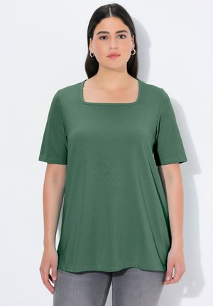 Frau mit glattem dunklem Haar, die goldene Creolen, ein grünes kurzärmeliges Oberteil mit quadratischem Ausschnitt und hellgraue Hose trägt, steht an einer weißen Wand.