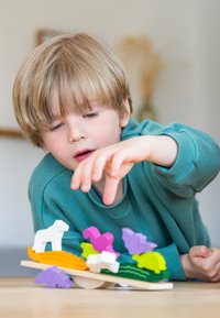 Kleurrijk houten balansspeelgoed met dierfiguren in wit, paars en groen. Kind dat zich uitstrekt om stukjes op een lichte houten basis te plaatsen.
