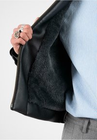 Czarna skórzana kurtka z miękkim, teksturowanym czarnym futrem wewnątrz, szczegółowy zamek błyskawiczny oraz ręka trzymająca otwartą kurtkę. Widoczna koszula w paski.