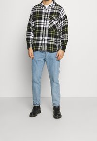 Camicia di flanella a quadri in bianco e nero con accenti verdi, chiusura a bottoni e tasca sul petto, abbinata a jeans azzurri chiari e stivali neri alla caviglia.