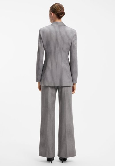 Femme avec les cheveux en chignon portant un costume gris à fines rayures, debout de dos, montrant le dos du blazer et du pantalon à jambes larges.