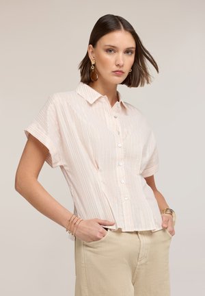 Frau mit kurzen braunen Haaren trägt eine hell gestreifte, kurzärmelige Bluse mit Knöpfen, beige Hose, goldene Ohrringe und mehrere goldene Armbänder.