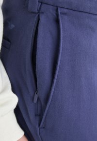 Pantaloni bleumarin dintr-un material texturat, dotati cu un buzunar cu fermoar și o croială adaptată, cu un talie netedă și detalii minime.