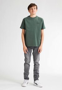 Zelené bavlněné tričko s kulatým výstřihem a krátkými rukávy, doplněné šedými džínami. Na hrudi je minimalistický textový detail. Tenisky jsou bílé.