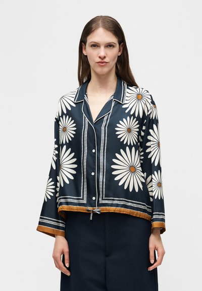 Γυναίκα που φοράει μπλε σκούρο πουκάμισο με κουμπιά, με μεγάλα λευκά σχέδια μαργαρίτας και μουσταρδί περίγραμμα, σε συνδυασμό με σκούρο παντελόνι, στέκεται μπροστά σε λευκό φόντο.