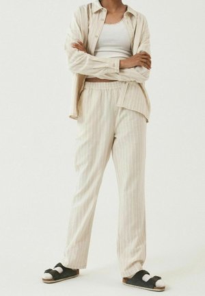 Personne vêtue d'un pantalon beige à rayures et d'une chemise assortie par-dessus un débardeur blanc, se tenant bras croisés, des sandales noires avec des chaussettes blanches.