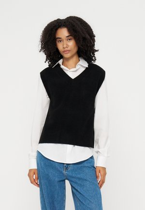 Femme aux cheveux bouclés portant un pull noir sans manches à col en V par-dessus une chemise blanche à col et un jean bleu sur un fond uni.
