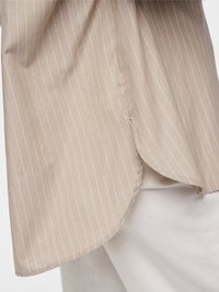 Šviesiai ruda dryžuota marškinėliai iš minkštos medžiagos su apvaliu apačios kraštu ir šoniniu plyšiu, dėvimi virš baltų, lygaus audinio kelnių.