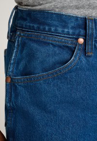 Blugi din denim albastru cu o textură netedă, nasturi din cupru și cusături vizibile. Detaliul buzunarului este parțial vizibil de-a lungul taliei.