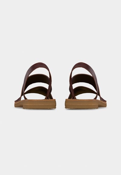 Ζευγάρι καφέ δερμάτινα σανδάλια με επίπεδες ταμπά σόλες, εμφανιζόμενα από πίσω σε ένα απλό ανοιχτό γκρι φόντο.