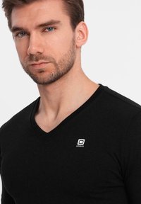 Černé tričko s výstřihem do V a přiléhavým střihem, vyrobené z měkké látky. Na levé části hrudi má malé bílé logo ve stylu "ombre".