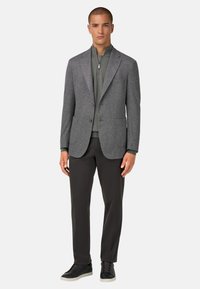 Šedá blazer s herringbone vzorem a klopovými límci, se dvěma předními kapsami, nošený přes zelenou košili na zip a doplněný o tmavé kalhoty a tenisky.