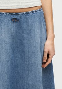 Κοντινό πλάνο ενός ατόμου που φοράει μπλε τζιν φούστα με ψηλή μέση, με ένα μικρό λογότυπο κοπής κοντά στη μέση και γκρι τοπ.