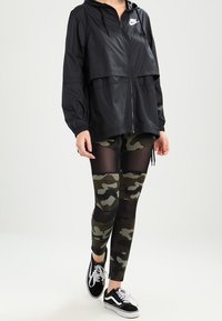 Svart vindjacka med huva och logotyp, ihop med svarta och gröna kamouflageleggings med nätpaneler, avslutat med svarta sneakers.