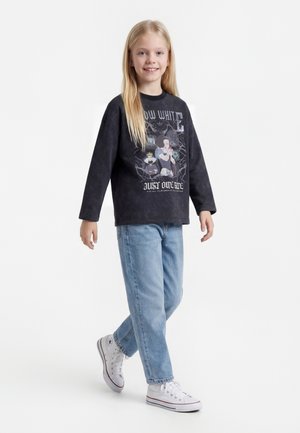 Fille aux longs cheveux blonds portant un sweat-shirt graphique Blanche-Neige noir, un jean bleu clair et des baskets blanches, marchant avec un sourire.