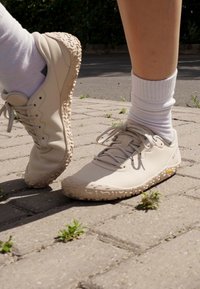 Beige sneakers with textured rubber soles, round toe, and lace-up design. White cotton socks visible, standing on a paved surface with greenery.