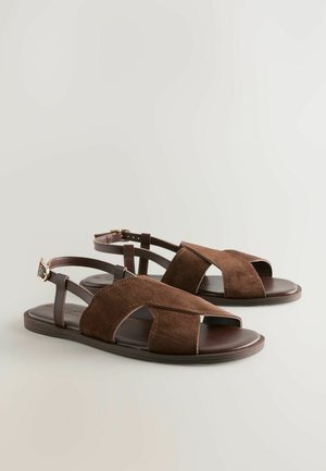 Sandale din suede maron cu talpa plată, prezentând bretele încrucișate și o cataramă ajustabilă la glezna. Design elegant cu accente decupate.