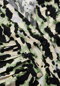 Button-up-Oberteil mit einem grünen und schwarzen Camouflage-Muster auf einem hellbeigen Hintergrund, mit gefältelten Details und schwarzen Knöpfen.