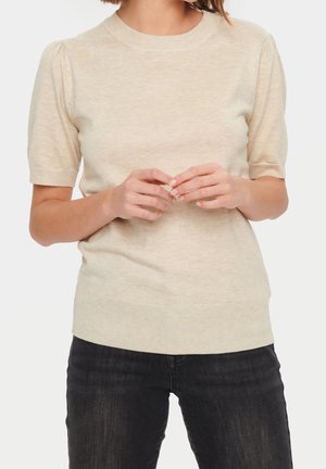 Vrouw draagt een beige gebreid shirt met korte mouwen en opgepofte schouders, gecombineerd met zwarte spijkerbroek, staand met handen losjes gevouwen.