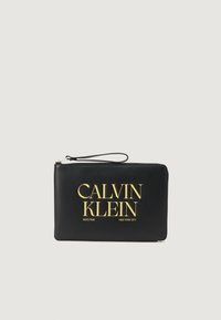 Sort læderhåndtaske med fremtrædende gul "CALVIN KLEIN" logo. Har en lynlås og håndledsrem for bekvemmelighed. Glat tekstur.