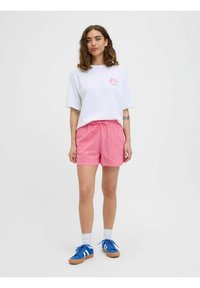 Balta medvilninė marškinė su maža rožine grafika, derinama su rožiniais trumpais šortais, papuoštais skylučių raštu. Mėlyni sportiniai bateliai ir balti kojinės.