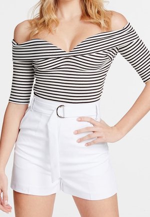 Vrouw die een zwart-wit gestreepte off-shoulder top draagt met ellebooglange mouwen en een witte short met hoge taille en een brede riem.