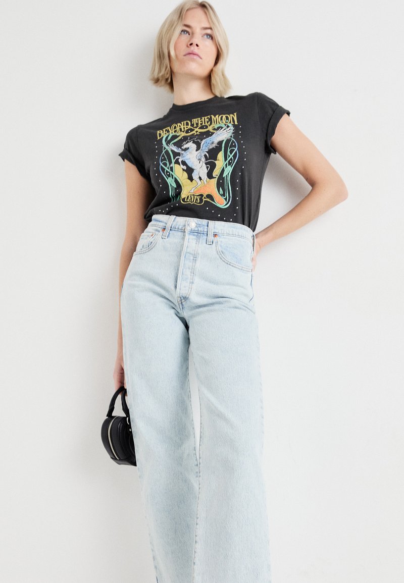 Camiseta gráfica negra con un diseño de "Más Allá de la Luna", combinada con jeans de talle alto en azul claro. Modelo sosteniendo un pequeño bolso negro.