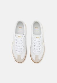 Adidași albi cu accente de piele întoarsă în bej, având o talpă din cauciuc și șireturi albe. Logo-ul auriu "BOSS" pe lateral și pe limba adidașilor.