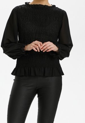 Kobieta ubrana w czarną, teksturowaną bluzkę z długim rękawem, z falbankami na mankietach i na dole, w połączeniu z czarnymi dopasowanymi spodniami, ręce złączone z przodu.