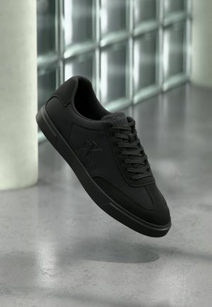 Svart lavt sneakers med snøring og "CK"-logo på siden, svevende over polert betonggulv nær glassblokkvegg.