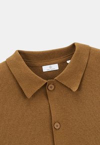 Polo en maille marron avec un tissu texturé, un col plat et deux boutons sur le devant. Étiquette indiquant "Serge Blanco."