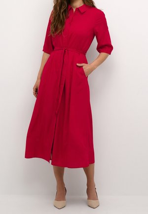 Femme portant une robe-chemise midi rouge avec les manches retroussées jusqu'aux coudes, une ceinture à la taille, des poches latérales, et des escarpins beige à bout pointu, debout contre un mur blanc.