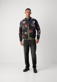 Μαύρο μπουφάν bomber με πολύχρωμα floral σχέδια, πράσινη πλέξη από κάτω, σκούρο τζιν και μαύρα παπούτσια, στέκεται σε ουδέτερο φόντο.