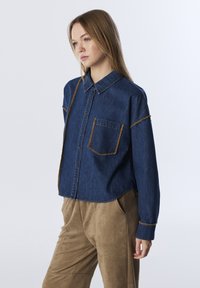 Donkerblauwe spijkerblouse met een patch pocket, koperkleurige stiksels en een kraag. Gecombineerd met beige, gestructureerde broek.