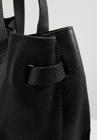 Gros plan sur le coin d'un sac à main en cuir noir texturé avec une lanière passée à travers une découpe rectangulaire.