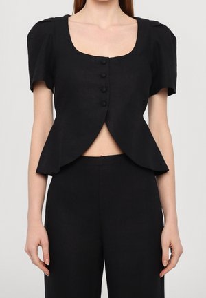 Vrouw draagt een zwart kortärmelig topje met vier knopen aan de voorkant en een uitlopende, open zoom, gecombineerd met zwarte broek met hoge taille.