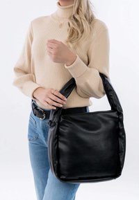 Mustassa nahkaisessa kassissa on yksi kaareva kahva. Sen pinta on sileä, muoto on suorakulmainen ja yläosa vetoketjullinen. Asusteena beige neule.